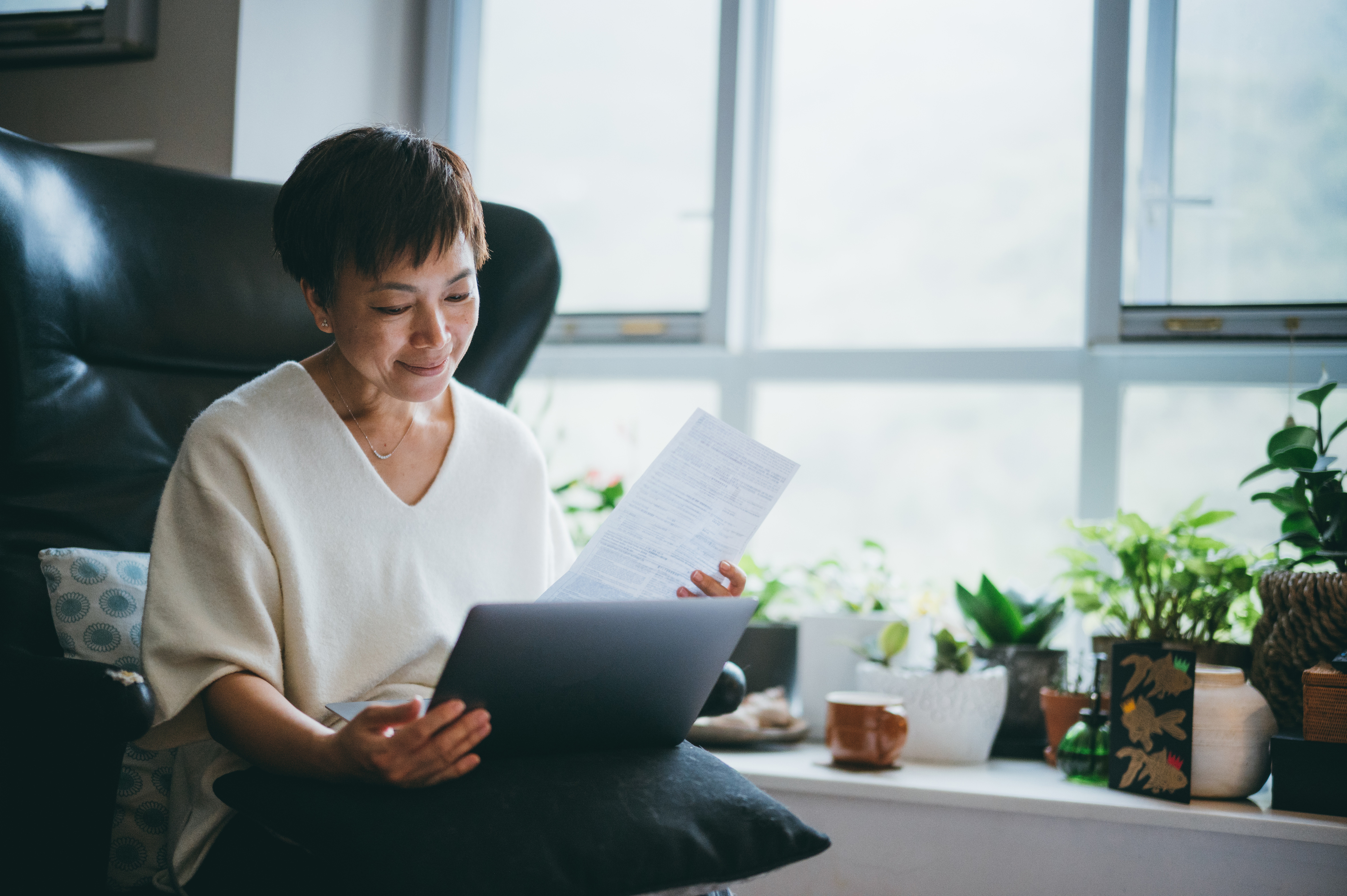 An older woman smiles at her laptop and some papers.